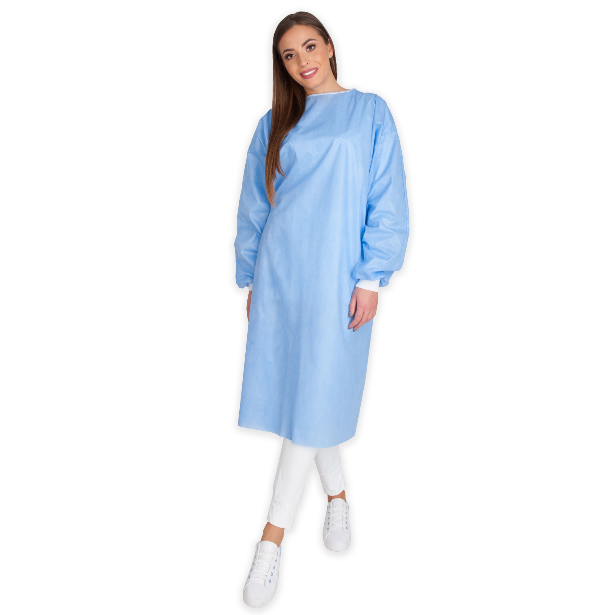 Uśmiechnięta kobieta prezentuje jasnoniebieski, jednorazowy fartuch medyczny z długimi rękawami i białymi mankietami, ubrana w białe spodnie i sportowe buty na białym tle.