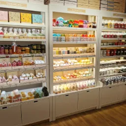 Ekspozycja w sklepie z produktami do pielęgnacji i aromaterapii: mydła glicerynowe, kule do kąpieli, świece zapachowe i zestawy prezentowe, ułożone na półkach w białych regałach z podświetleniem.