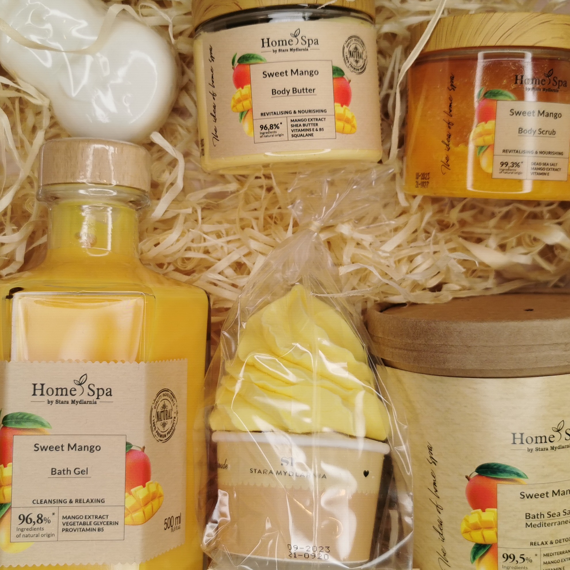 Zestaw prezentowy z kosmetykami Home Spa Sweet Mango: żel do kąpieli, masło do ciała, peeling do ciała i mydełko w kształcie deseru, ułożone na dekoracyjnej słomie.