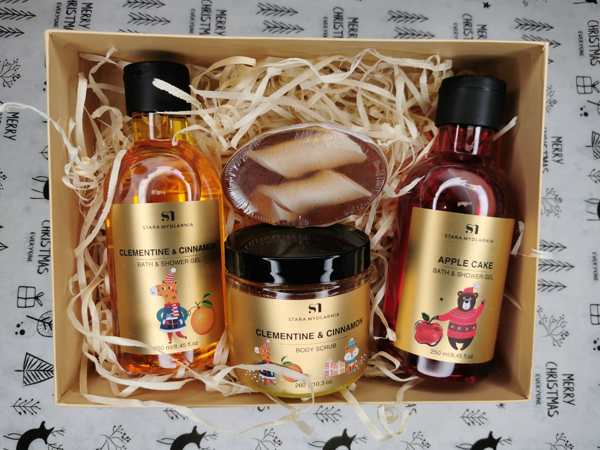 Świąteczny kosz prezentowy z naturalnymi kosmetykami Stara Mydlarnia: żel do kąpieli o zapachu clementiny i cynamonu, żel do kąpieli o zapachu szarlotki oraz peeling do ciała, ułożone...