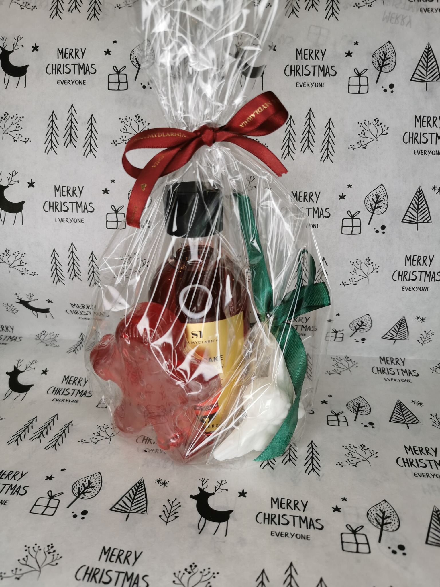 Świąteczny kosz prezentowy z mydłem w kształcie gwiazdy, olejkiem i musującą kulą do kąpieli, zapakowany w folię celofanową z czerwoną wstążką z napisem i zieloną wstążką, na papierze z motywem...