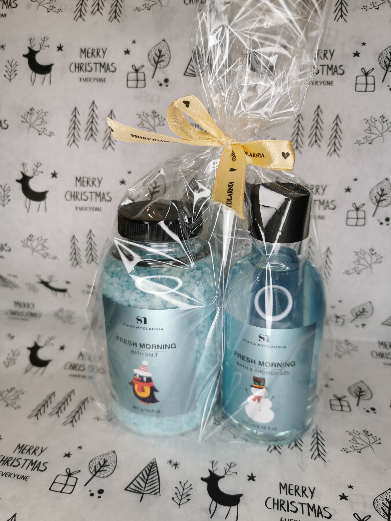 Dwa turkusowe pojemniki z kosmetykami Stara Mydlarnia, sól do kąpieli i żel pod prysznic, zapakowane w przezroczysty celofan i przewiązane kremową wstążką z nadrukiem nazwy firmy, na tle papieru...