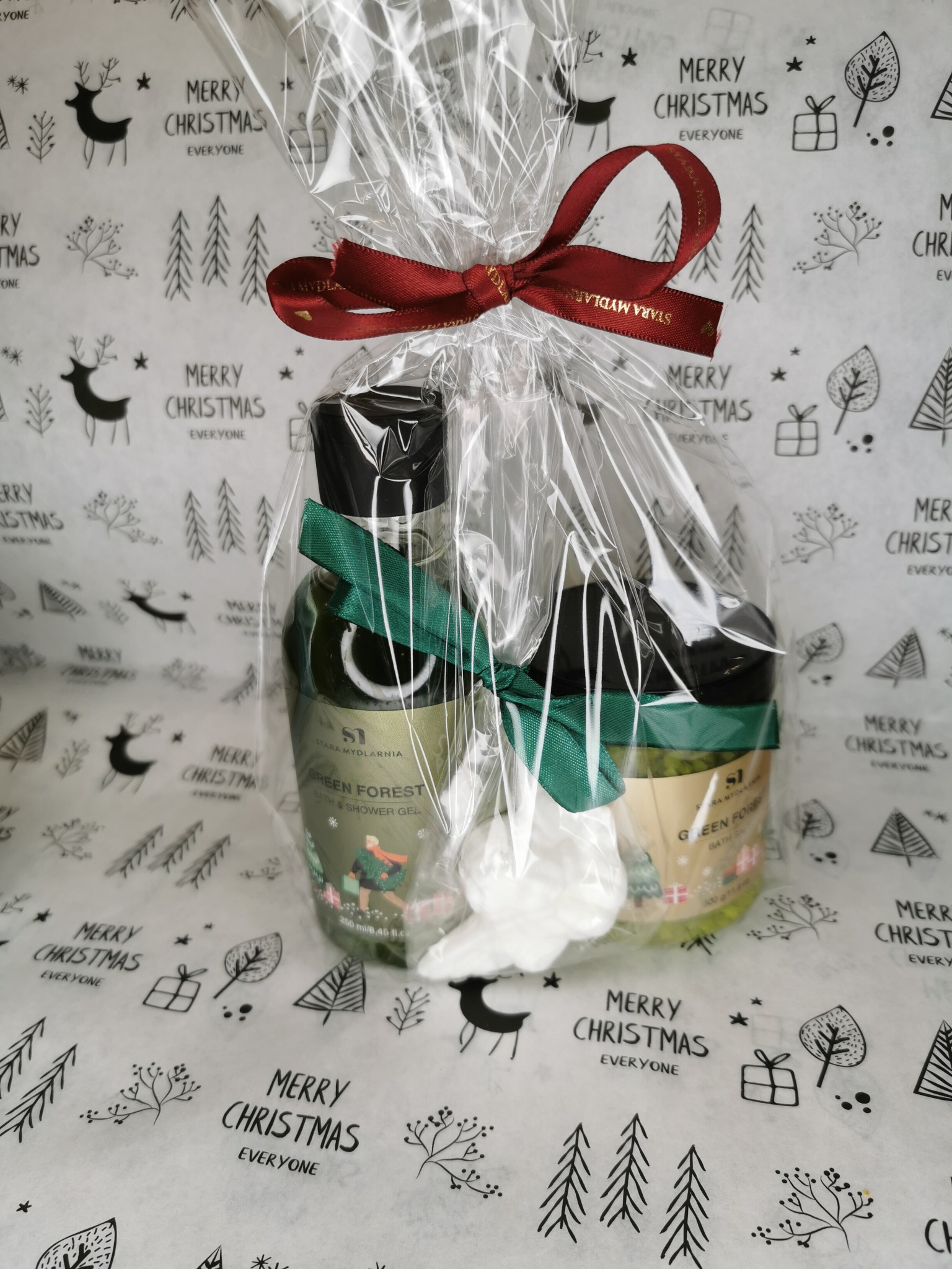 Świąteczny kosz prezentowy z kosmetykami marki Stara Mydlarnia, zapakowany w przezroczystą folię z czerwoną kokardą na tle papieru z motywem świątecznym.