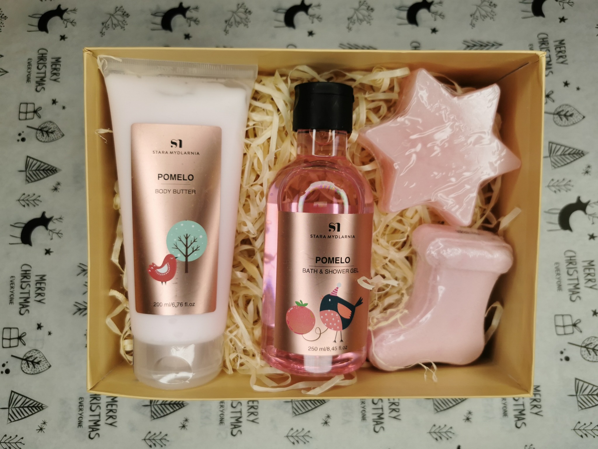 Beżowy kosz prezentowy wypełniony kremem do ciała, żelem pod prysznic i dwoma mydełkami w kształcie gwiazdy i buta, wszystko marki Stara Mydlarnia, na tle papieru w świąteczne wzory.