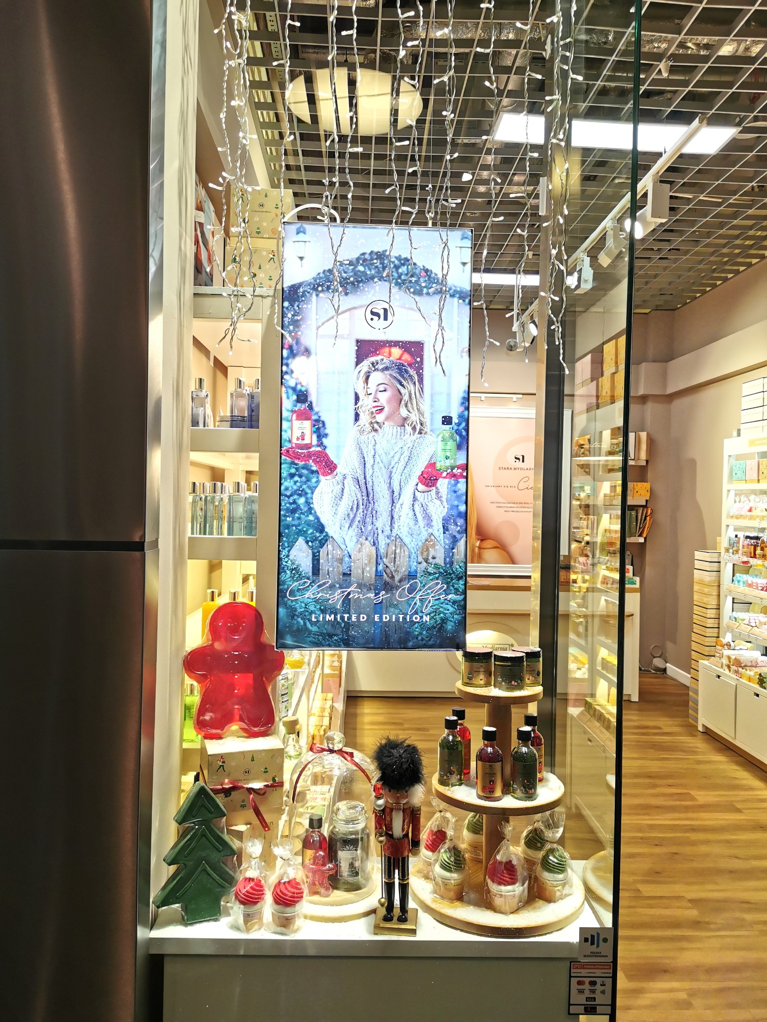 Świąteczna witryna sklepowa z kosmetykami i ozdobami, w tym piernikowy ludzik z mydła, drewniany dziadek do orzechów i opakowane babeczki z kremem, dekorowana lampkami.