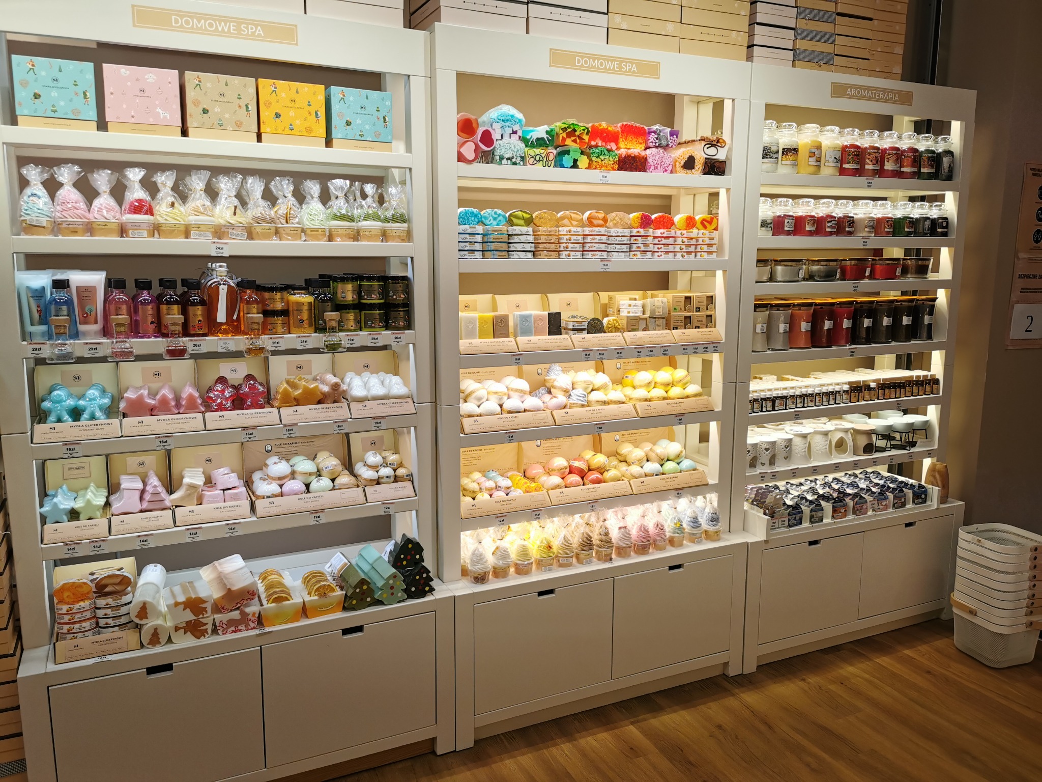 Ekspozycja w sklepie z produktami do pielęgnacji i aromaterapii: mydła glicerynowe, kule do kąpieli, świece zapachowe i zestawy prezentowe, ułożone na półkach w białych regałach z podświetleniem.