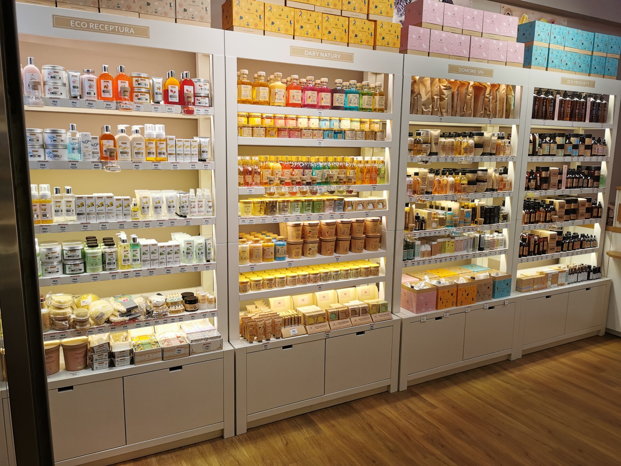Półki sklepowe wypełnione różnorodnymi produktami kosmetycznymi i do pielęgnacji, ułożonymi w estetyczny sposób, z podziałem na kategorie oznaczone napisami 'Eco Receptura', 'Dary Natury', 'Domowe...