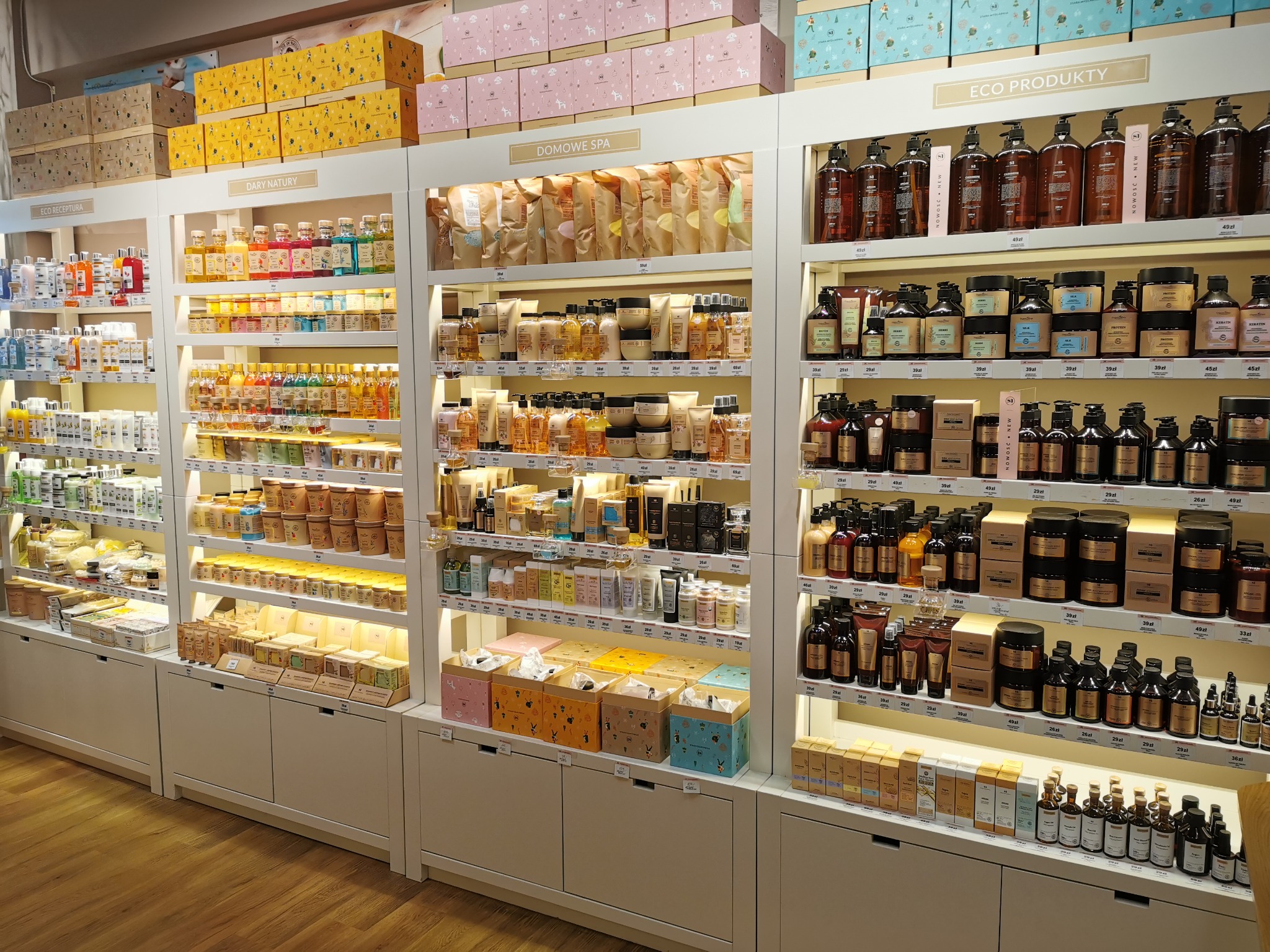 Półki sklepowe z szerokim asortymentem kosmetyków naturalnych i produktów do pielęgnacji, w estetycznych opakowaniach i kartonach prezentowych, podświetlone ciepłym światłem.