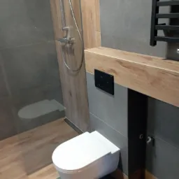 Nowoczesna łazienka z szarymi płytkami, drewnopodobną zabudową, wiszącą toaletą i czarnym grzejnikiem drabinkowym.