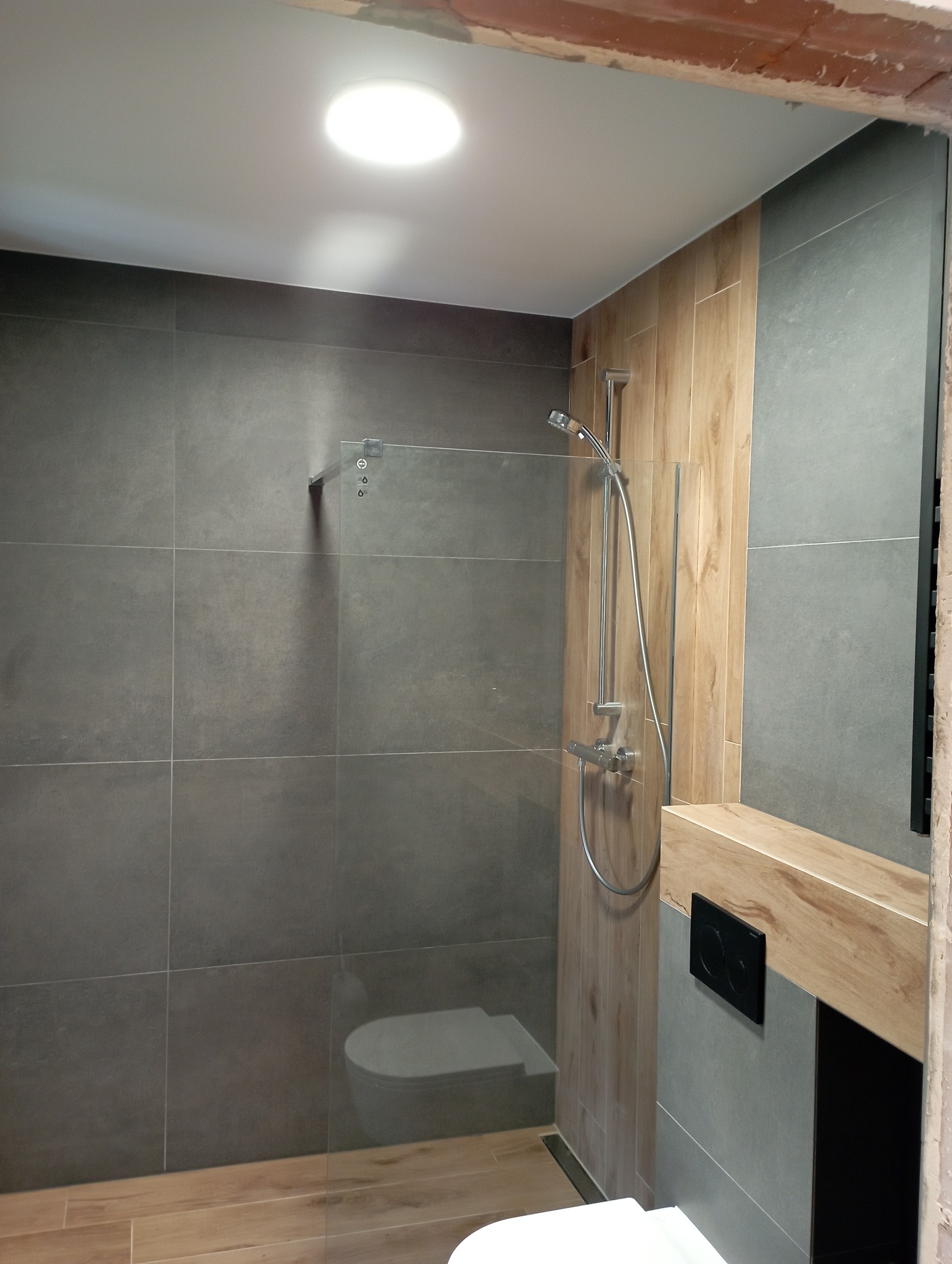 Nowoczesna łazienka z kabiną prysznicową typu walk-in, szare płytki na ścianach i podłodze imitujące drewno, deszczownica oraz czarny panel sterowania toaletą podtynkową.