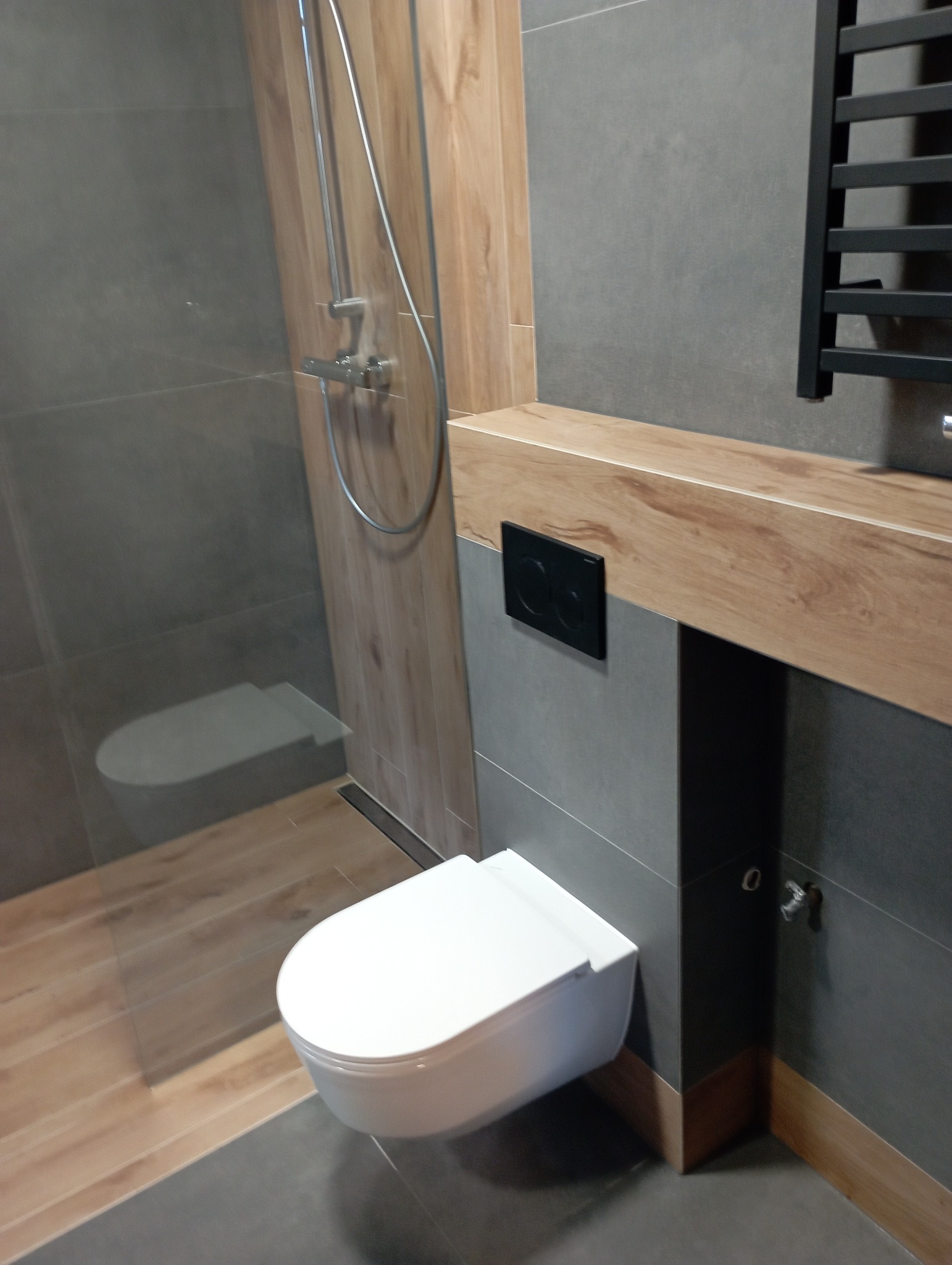 Nowoczesna łazienka z szarymi płytkami, drewnopodobną zabudową, wiszącą toaletą i czarnym grzejnikiem drabinkowym.
