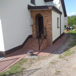 Wykończona elewacja domu jednorodzinnego z jasną fasadą, ciemnobrązowymi akcentami, wejściem wyłożonym cegłą i ścieżką z kostki; na trawniku widać dwóch pracowników.