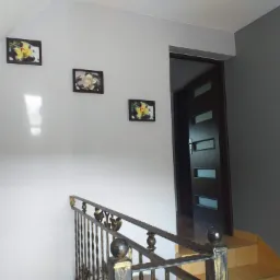 Korytarz na poddaszu z szarą ścianą, trzema obrazkami z motywem kwiatowym, ciemnymi drzwiami z przeszkleniami i balustradą schodów z metaloplastyki.