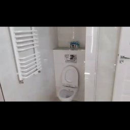 Kosmiczne Remonty - Biała, błyszcząca łazienka po remoncie, widoczna toaleta z otwartą klapą, grzejnik drabinkowy i częściowo widoczne drzwi, na półce nad toaletą leżą rolki papieru toaletowego.