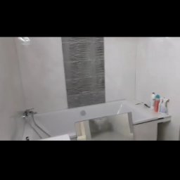 Kosmiczne Remonty - Wykończona łazienka z biało-szarymi płytkami, wanną z baterią i umywalką w zabudowie z blatem, na którym stoją kosmetyki.