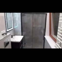 Nowoczesna łazienka z kabiną prysznicową o szarych ścianach, umywalką z ciemną szafką, podświetlanym lustrem i grzejnikiem drabinkowym przy oknie.