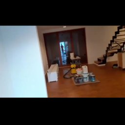 Kosmiczne Remonty - Wnętrze w trakcie przygotowań do malowania: widoczny odkurzacz, pojemniki z farbą i akcesoriami malarskimi na kartonowym podkładzie; w tle przeszklone drzwi i sylwetka osoby.