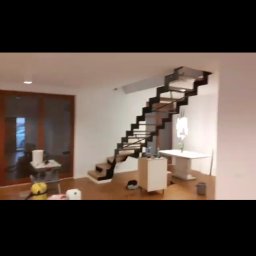 Kosmiczne Remonty - Wnętrze w trakcie prac: nowoczesne schody z czarnymi wspornikami i drewnianymi stopniami, widoczne narzędzia i akcesoria malarskie, częściowo umeblowane pomieszczenie z drewnianą podłogą.