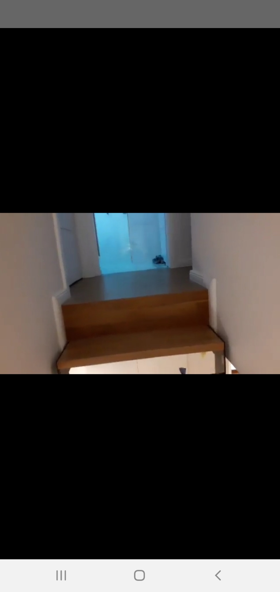 Widok na schody wewnętrzne z drewnianymi stopniami i białymi ścianami, prowadzące do pomieszczenia z jasną podłogą i niebieskim oświetleniem.