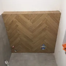 Ściana w łazience wyłożona brązowymi płytkami imitującymi drewno, ułożonymi w jodełkę, z widocznymi krzyżykami dystansowymi, instalacją wodną i szarymi płytkami na podłodze i sąsiedniej ścianie.