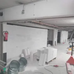 Wznoszenie ściany z białych bloczków w surowym wnętrzu, widoczne materiały budowlane, instalacje pod sufitem i gaśnica na ścianie