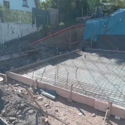 Zbrojenie fundamentu domu jednorodzinnego z widoczną siatką stalową, deskowaniem i elementami instalacji podposadzkowej na tle skarpy z rurami drenażowymi.