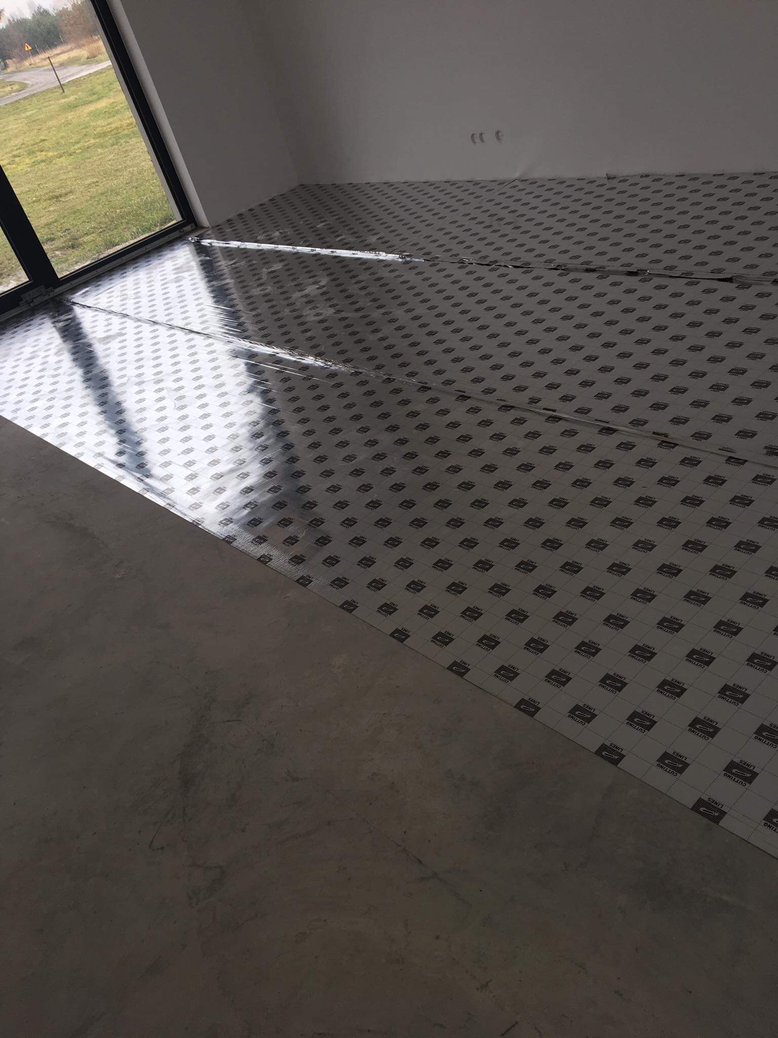 Przygotowanie podłoża pod płytki: rozłożona folia ochronna z nadrukiem na betonowej wylewce w nowym budynku, widok z okna.