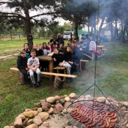 Grupa dzieci i dorosłych siedzi przy drewnianych stołach w plenerze, w tle las i budynek, na pierwszym planie grill z dużą ilością piekących się kiełbas, unosi się dym.