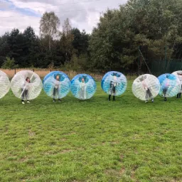 Grupa dzieci w przezroczystych i niebieskich kulach Bumper Ball na trawiastym boisku, w tle drzewa i siatka bramki.