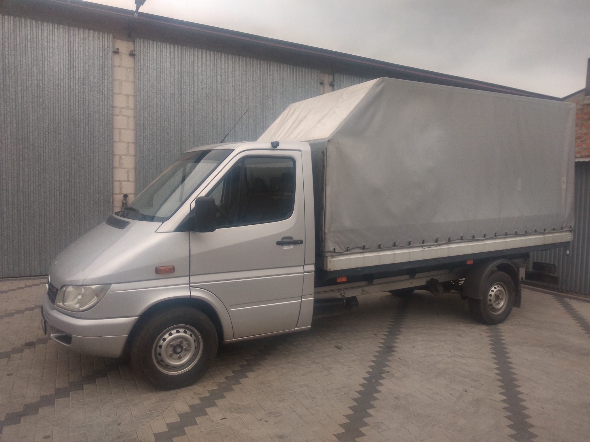 Srebrny samochód dostawczy typu plandeka zaparkowany na brukowanym podjeździe przed szarym metalowym garażem.