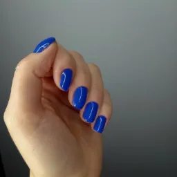 Dłoń z krótkimi, kwadratowymi paznokciami pomalowanymi na intensywny, kobaltowy odcień lakieru hybrydowego, widoczne delikatne refleksy światła na gładkiej powierzchni.