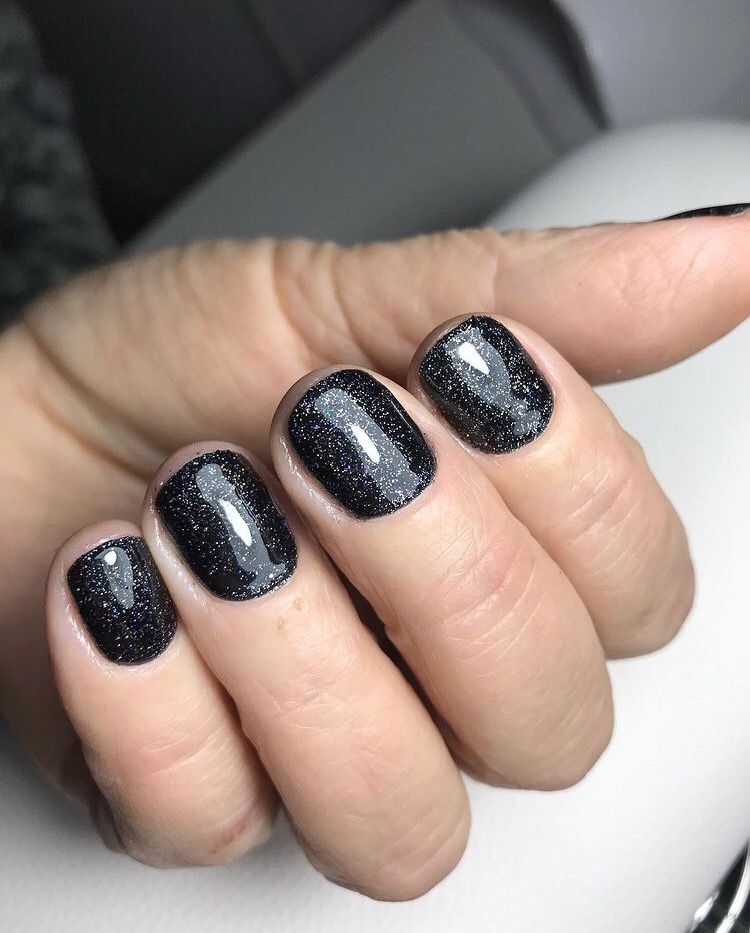 Czarny manicure hybrydowy z brokatem na krótkich, kwadratowych paznokciach, widoczne delikatne zmarszczki na skórze palców.