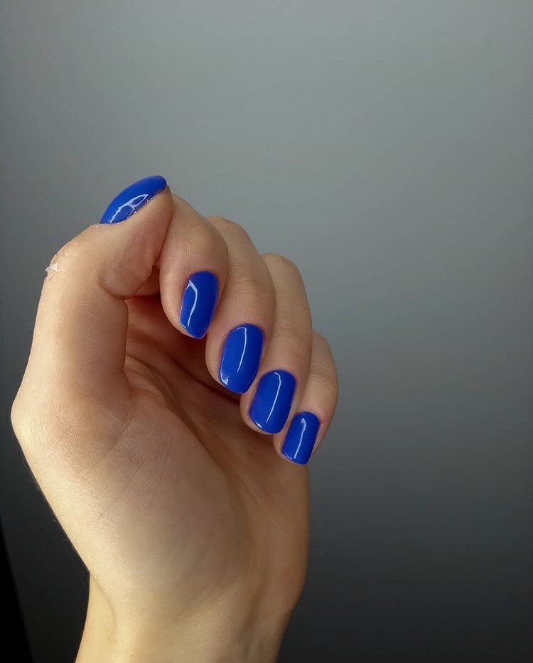 Dłoń z krótkimi, kwadratowymi paznokciami pomalowanymi na intensywny, kobaltowy odcień lakieru hybrydowego, widoczne delikatne refleksy światła na gładkiej powierzchni.