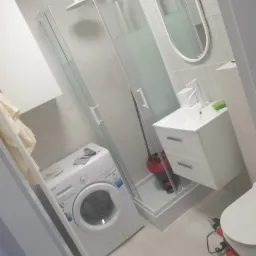 Wnętrze łazienki po remoncie: kabina prysznicowa z brodzikiem, pralka, umywalka z szafką, owalne lustro i narzędzia (wiertarka) na podłodze.