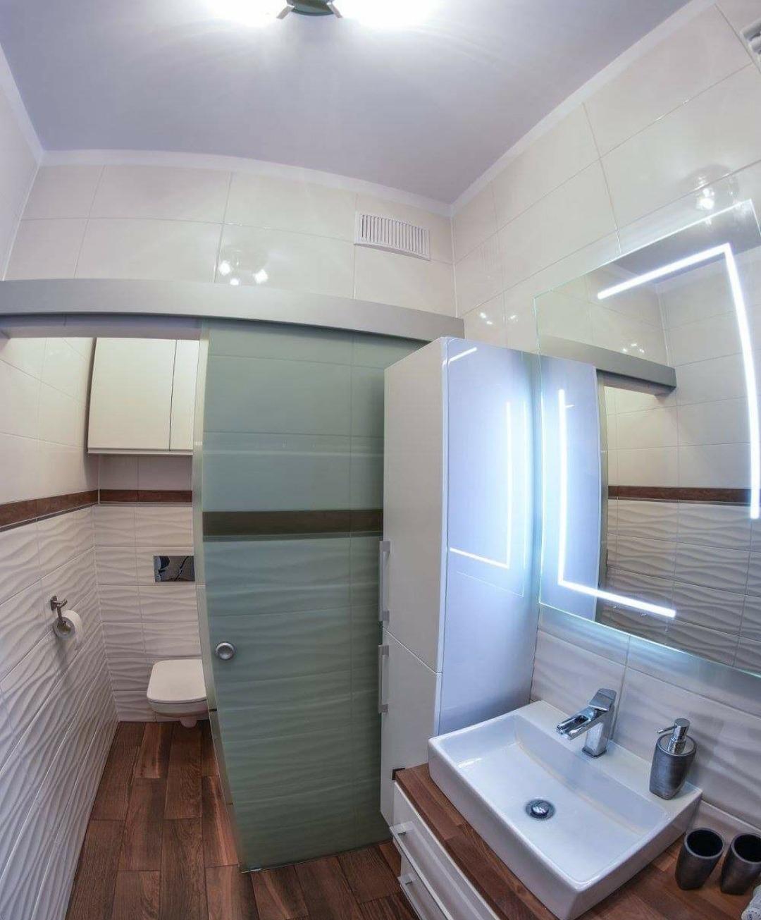 Nowoczesna łazienka z przesuwnymi drzwiami z matowego szkła, podświetlanym lustrem, białą umywalką nablatową na drewnianym blacie i toaletą w tle.