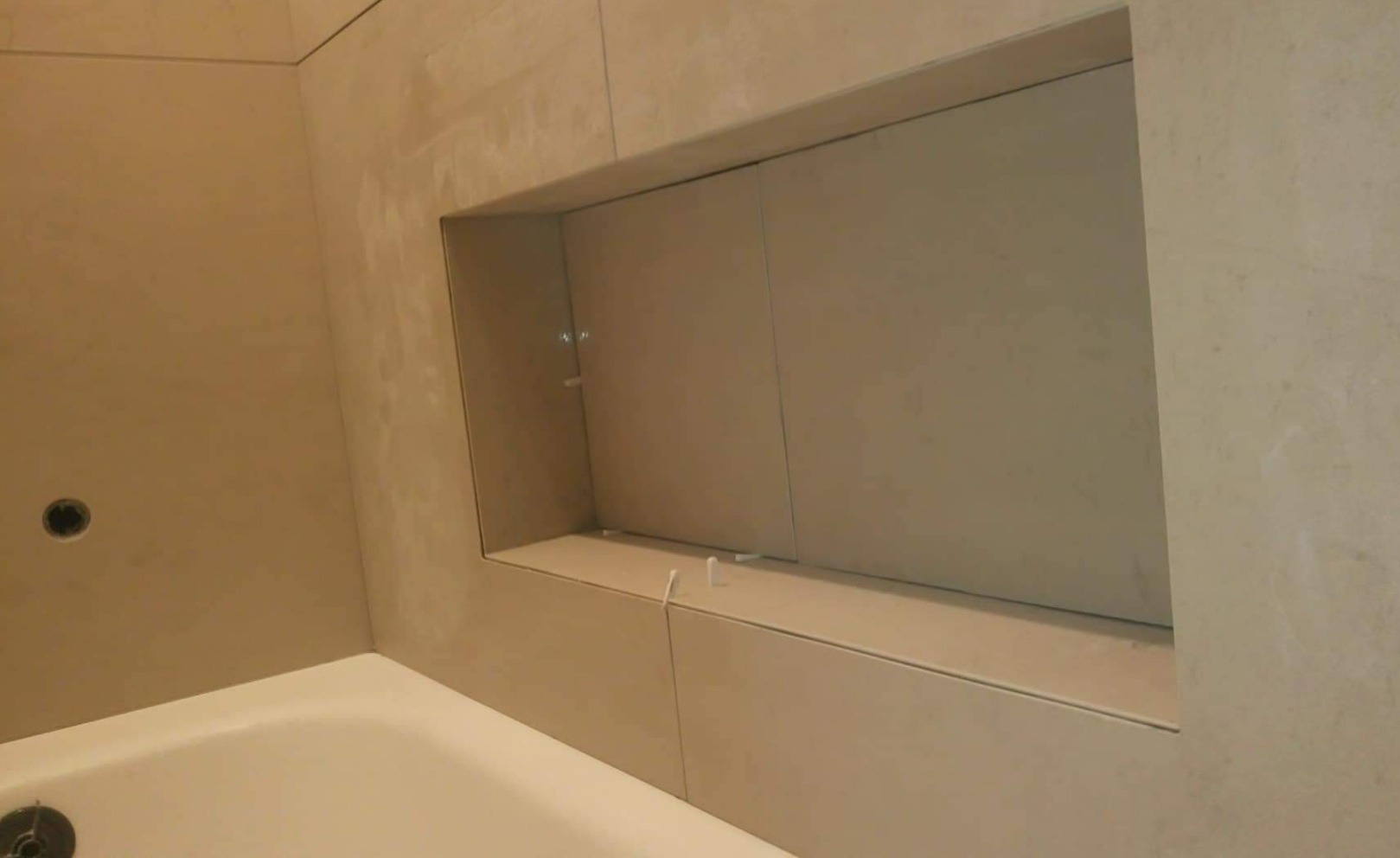 Narożnik łazienki z wanną i wnęką w ścianie wyłożoną płytkami w odcieniach beżu, widoczne elementy montażowe wewnątrz wnęki.