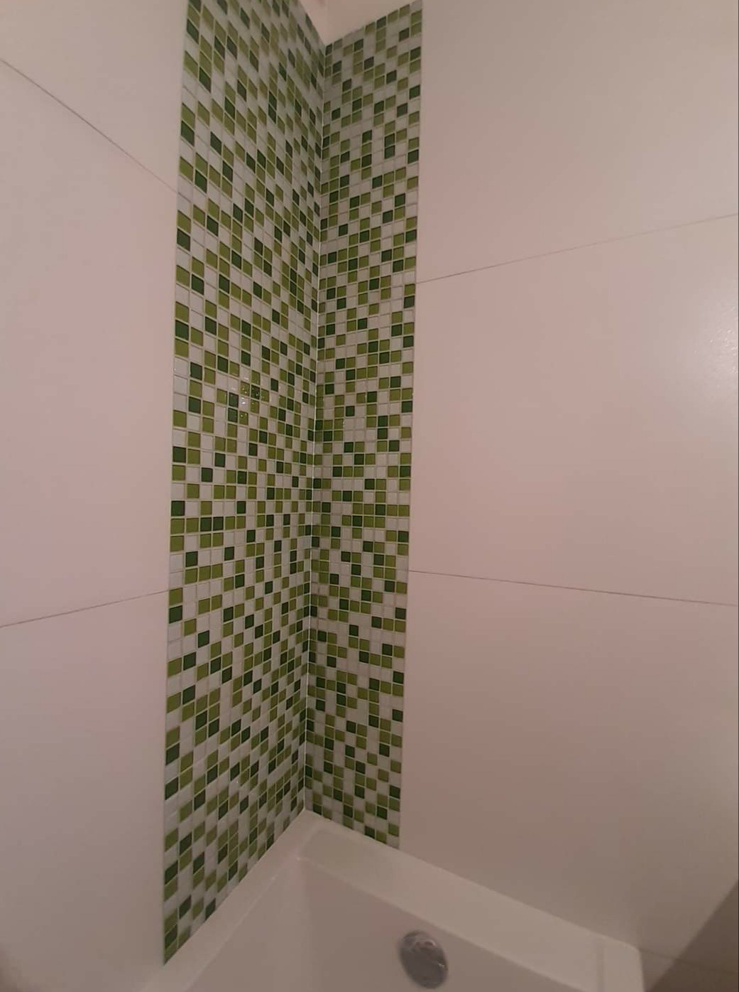 Fragment ściany łazienki z białymi płytkami i pionowym pasem mozaiki w odcieniach zieleni, widoczny fragment wanny.