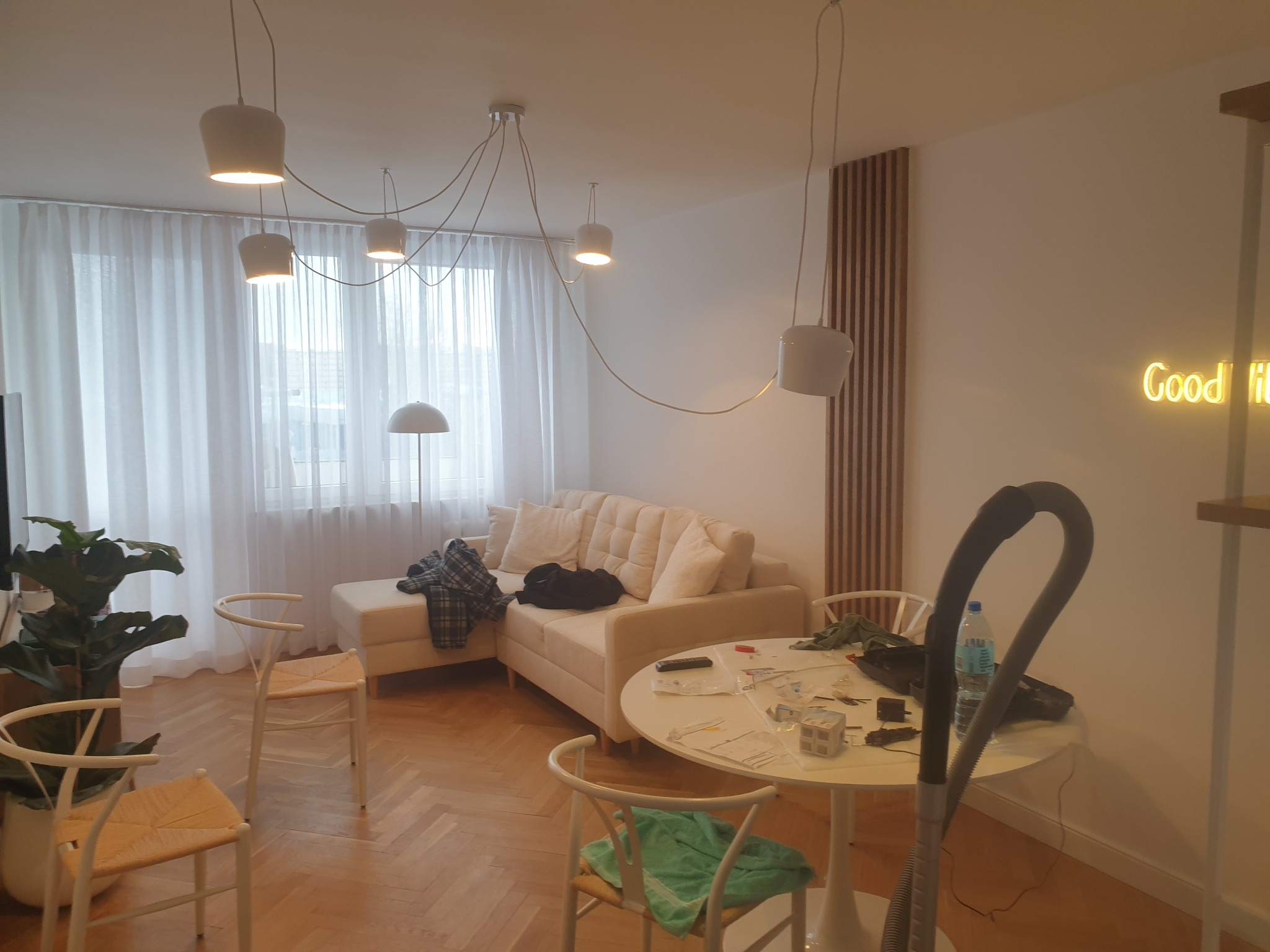 Salon w trakcie sprzątania: kremowa sofa z poduszkami, okrągły stół z akcesoriami, białe krzesła, lampa sufitowa z kilkoma kloszami na kablach, odkurzacz. Na ścianie dekoracyjny panel z drewnianych...