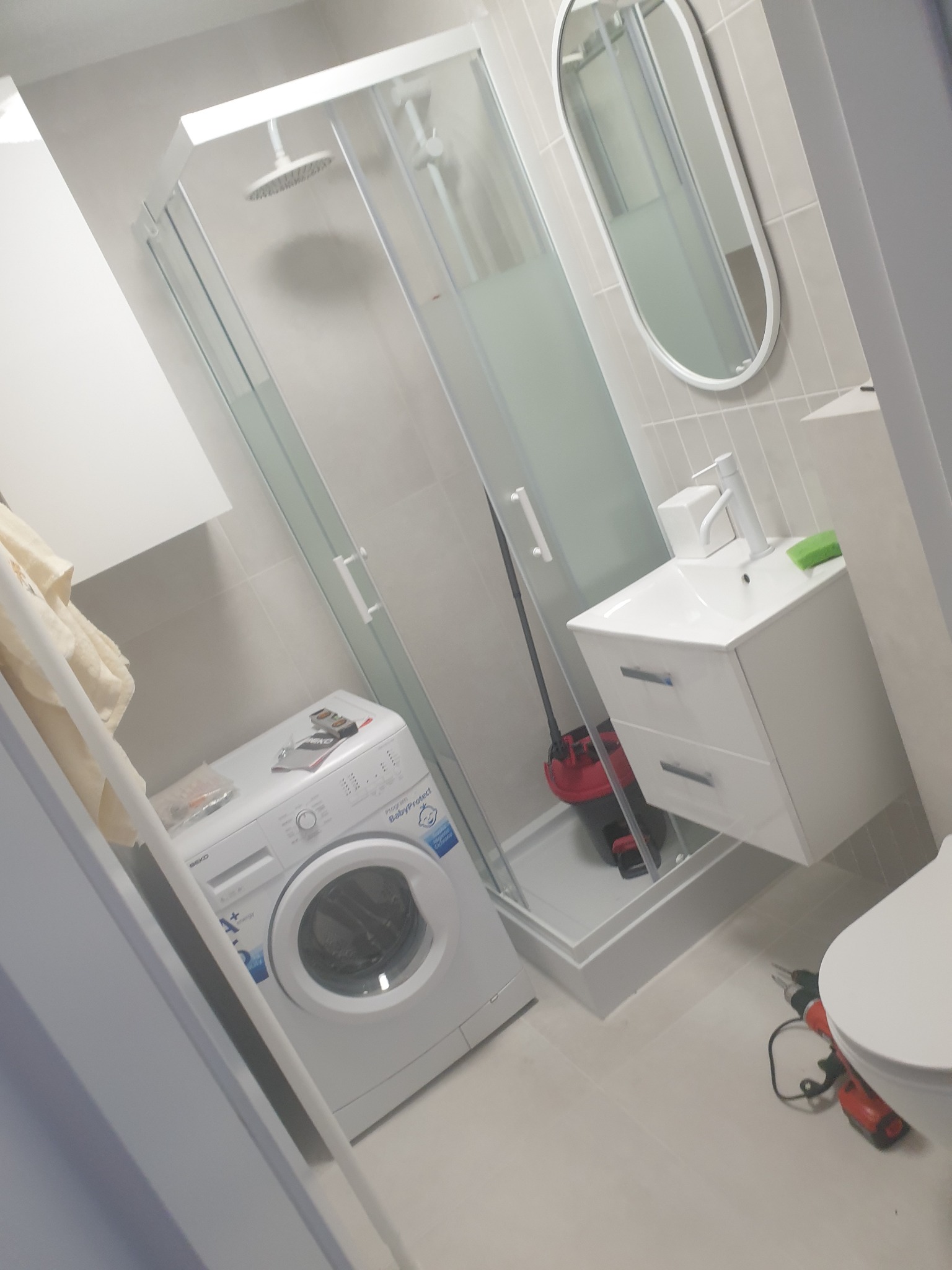 Wnętrze łazienki po remoncie: kabina prysznicowa z brodzikiem, pralka, umywalka z szafką, owalne lustro i narzędzia (wiertarka) na podłodze.