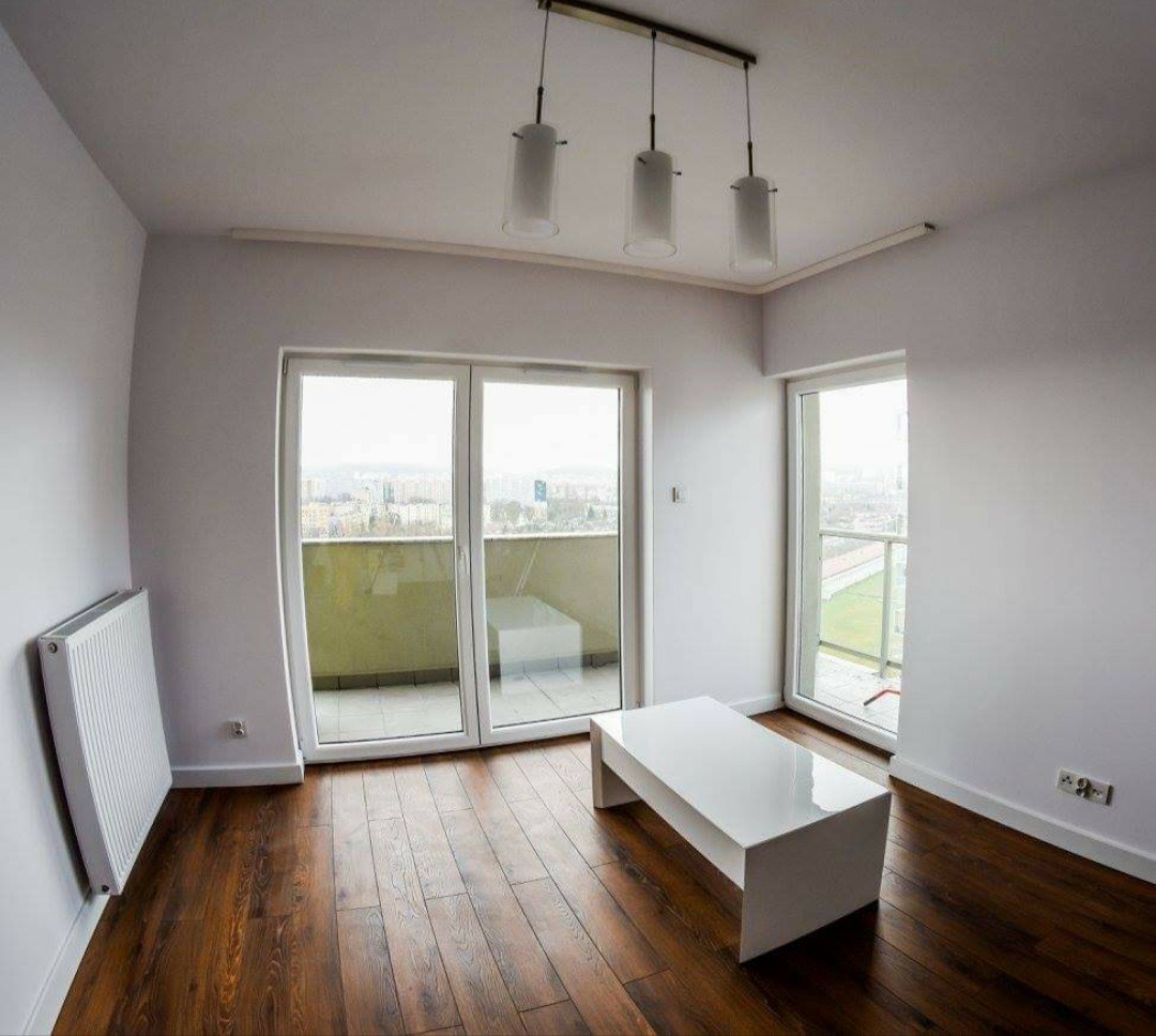 Pomalowane na biało wnętrze pokoju po remoncie w Gdańsku, z nową, błyszczącą podłogą z ciemnego drewna, białym grzejnikiem na ścianie, białym stolikiem kawowym i widokiem na miasto z balkonu.