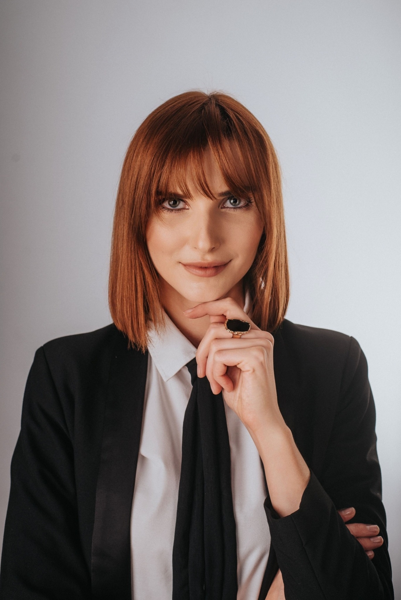 Portret kobiety z rudymi włosami w czarnym garniturze, z dłonią pod brodą i dużym pierścieniem, na jasnym tle. Profesjonalny wizerunek.