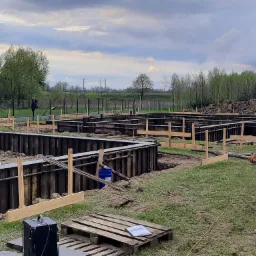 Przygotowanie szalunków pod fundamenty domu murowanego, widoczne metalowe ścianki szalunkowe i drewniane podpory, paleta z dokumentacją na pierwszym planie, krajobraz z drzewami w tle.