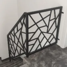 Nowoczesna, czarna balustrada schodowa z geometrycznym wzorem, widoczna na tle białej ściany i szarych schodów pokrytych wykładziną. W górnym lewym rogu widoczne lampy sufitowe o geometrycznym...