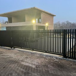 Usługi Spawalnicze Spaw-Mag - Nowoczesna, czarna brama przesuwna z pionowych profili na tle surowego budynku w trakcie budowy, oświetlonego lampami. Widoczne ślady opon na ziemi.