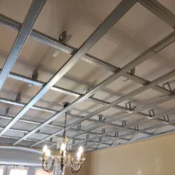 Konstrukcja sufitu podwieszanego z profili metalowych, widoczne elementy mocujące, w tle fragment żyrandola kryształowego i ściana w kolorze beżowym.