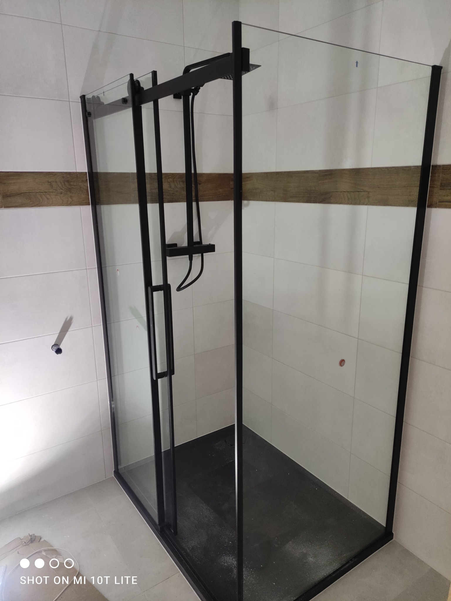 Kabina prysznicowa z czarnymi profilami i deszczownicą, zamontowana w łazience z jasnymi płytkami i dekoracyjnym pasem z imitacji drewna.