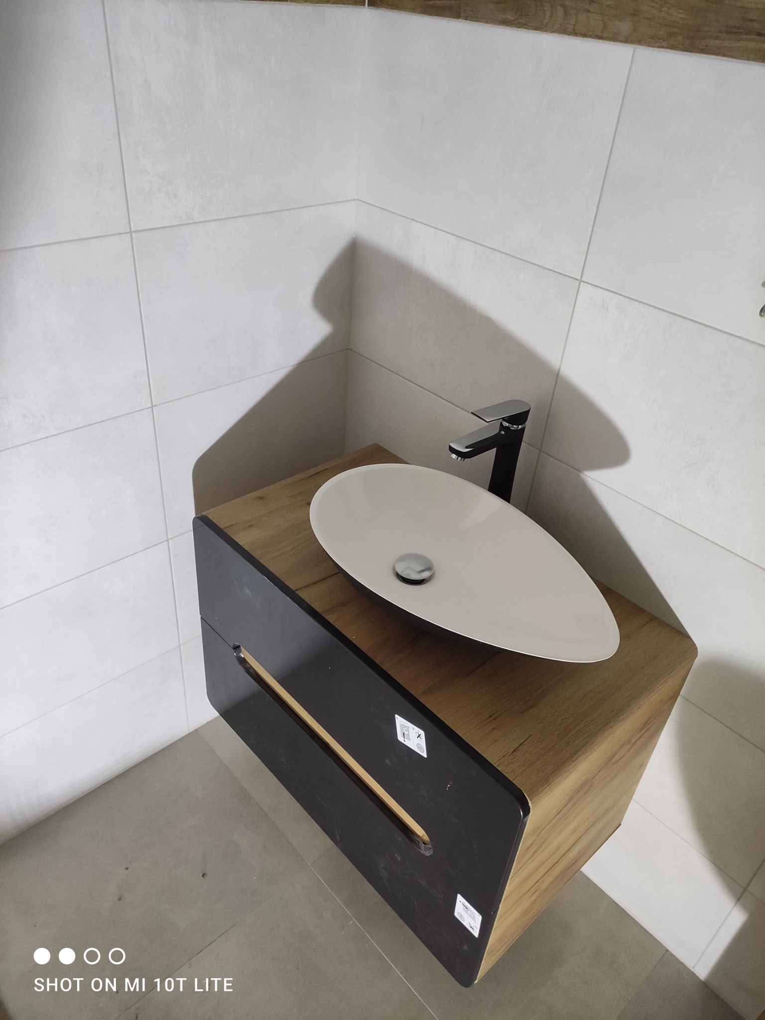 Nowoczesna łazienka z owalną białą umywalką na szafce z drewnopodobnym blatem i czarnym frontem, minimalistyczna bateria, jasne płytki na ścianach i podłodze.