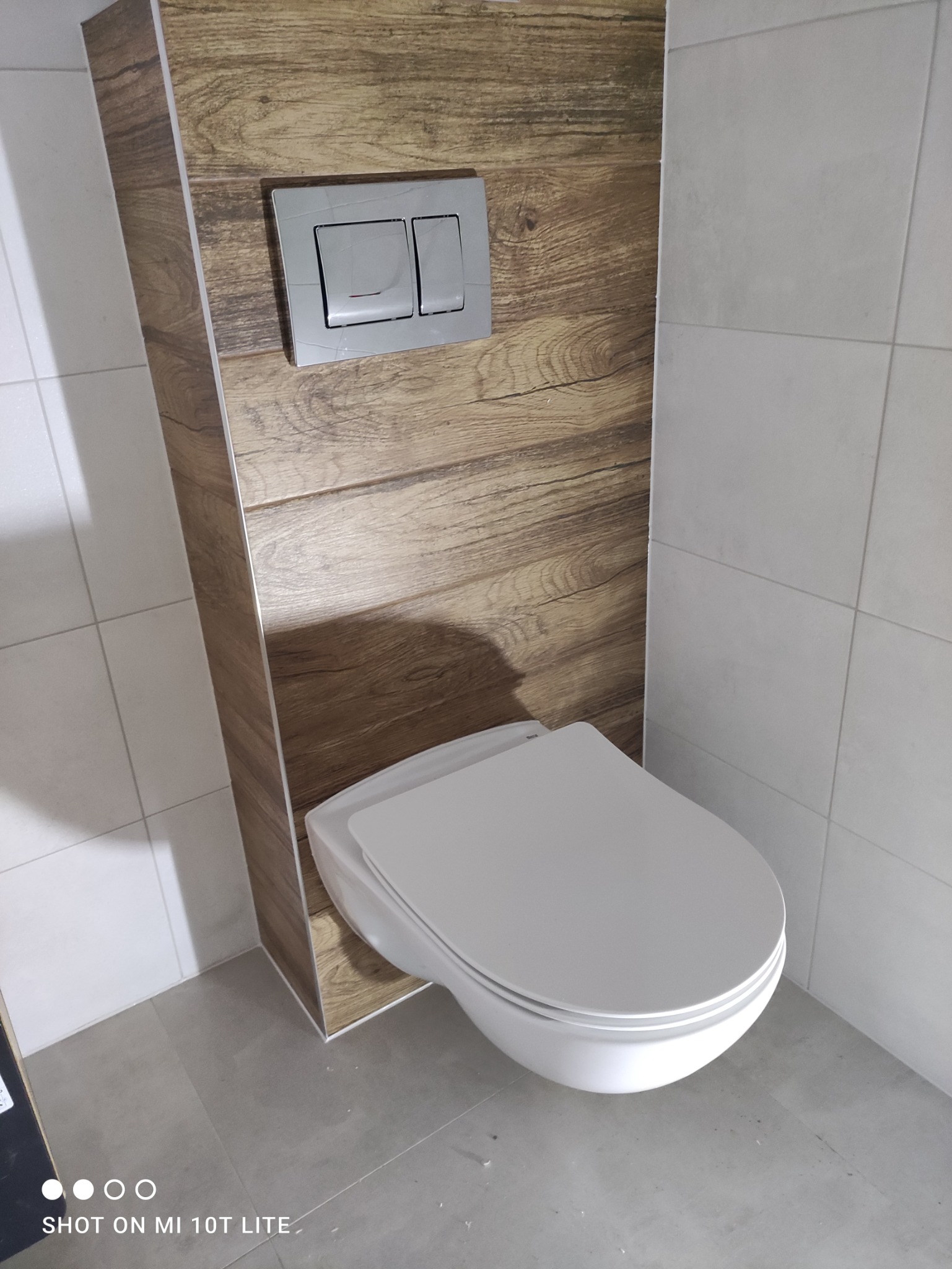 Zabudowa WC z drewnopodobną okładziną i przyciskiem spłukującym w szarym odcieniu, widoczne płytki ceramiczne na ścianach i podłodze.