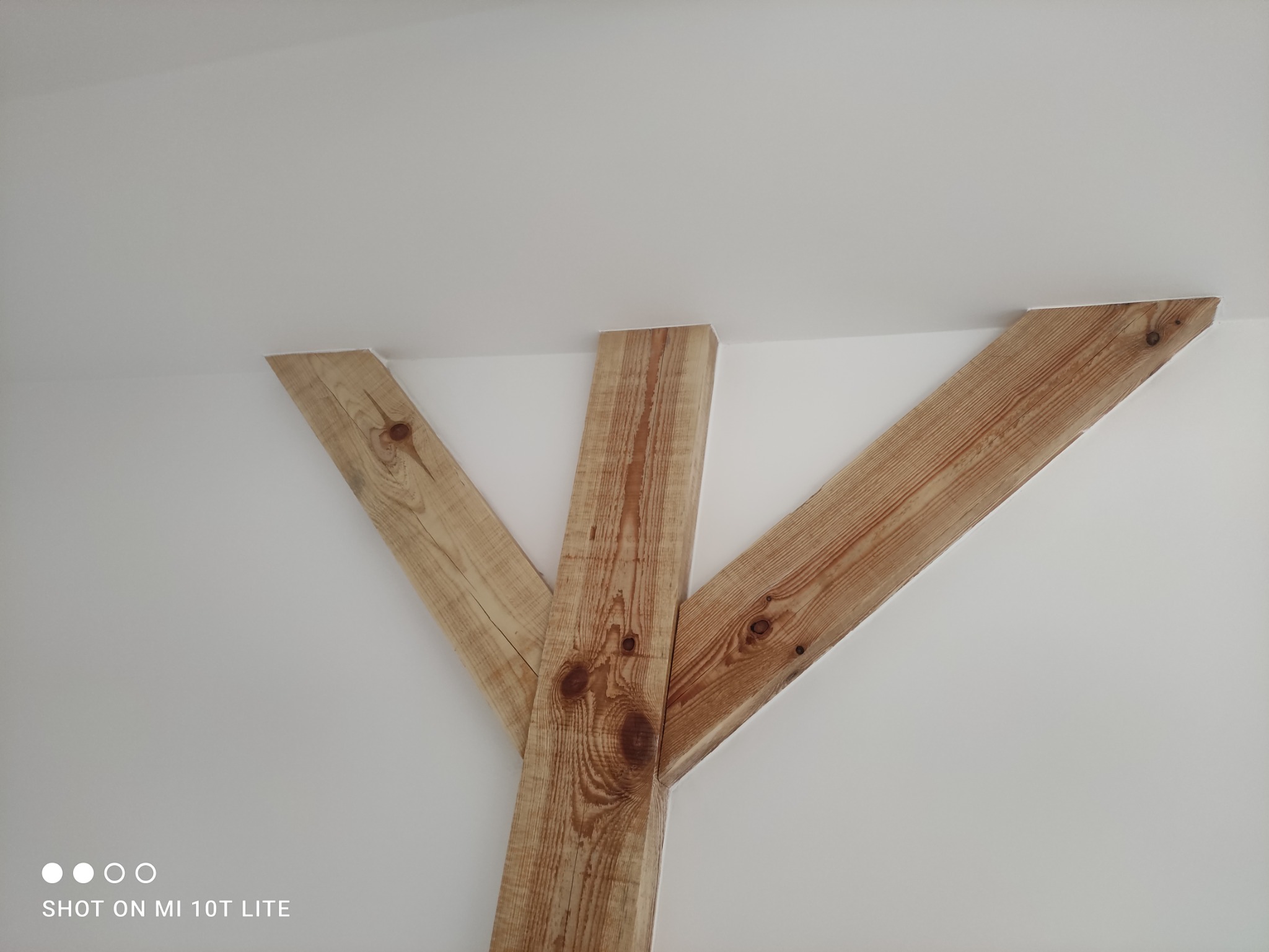 Drewniane belki dekoracyjne w kształcie litery Y, zamontowane przy suficie i ścianie w białym pomieszczeniu.