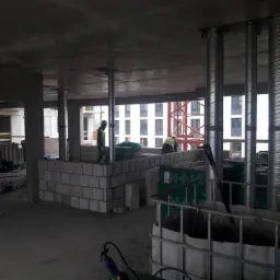 Wnętrze surowego budynku w trakcie budowy, widoczne murowane ściany z betonu komórkowego, stalowe podpory stropu, dwóch robotników w odblaskowych kamizelkach oraz pojemnik IBC z zaprawą.